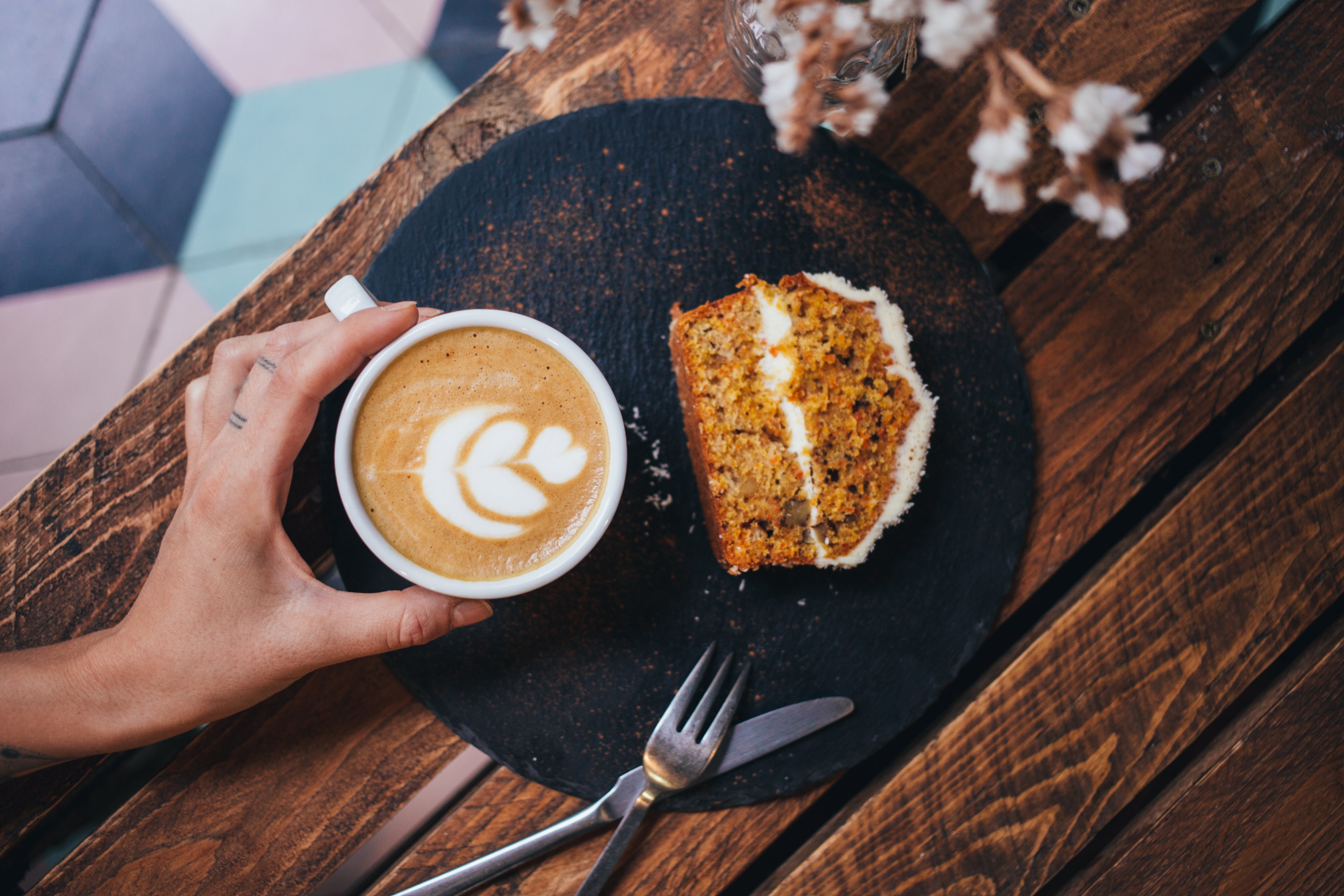 Hand hält eine cremige Tasse Cappuccino neben einem Stück Kuchen auf einem dunklen Keramikteller auf rustikalem Holztisch. Der aufgeschäumte Kaffee, die goldbraune Kuchenkruste und die warme Café-Atmosphäre vermitteln den Duft von frisch geröstetem Kaffee, süßem Gebäck und einen ruhigen Moment bewussten Genusses.