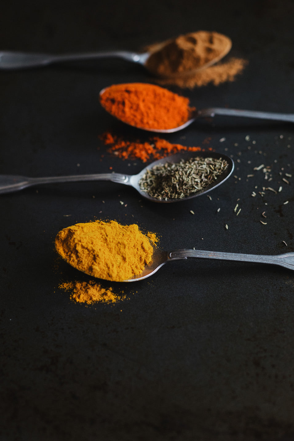 Mehrere Metalllöffel mit fein gemahlenen Gewürzen und Kräutern in warmen Gelb-, Rot- und Brauntönen auf dunkler Oberfläche. Die intensiven Farben, die pudrige Textur und die natürliche Präsentation vermitteln aromatische Vielfalt, erdige Gewürzdüfte und den Eindruck hochwertig ausgewählter Inhaltsstoffe.