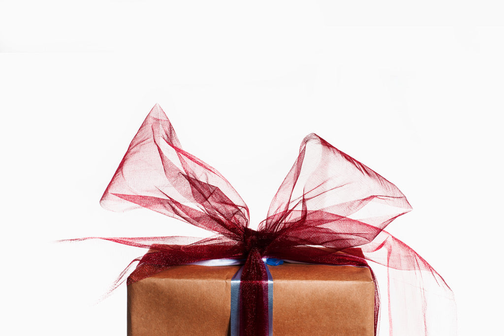 In braunes Papier eingepacktes Geschenk mit großer transparenter roter Schleife und dekorativem Band vor ruhigem hellem Hintergrund. Die warmen Farbtöne, die weichen Stoffstrukturen und die elegante Geschenkverpackung vermitteln eine festliche und hochwertige Atmosphäre.
