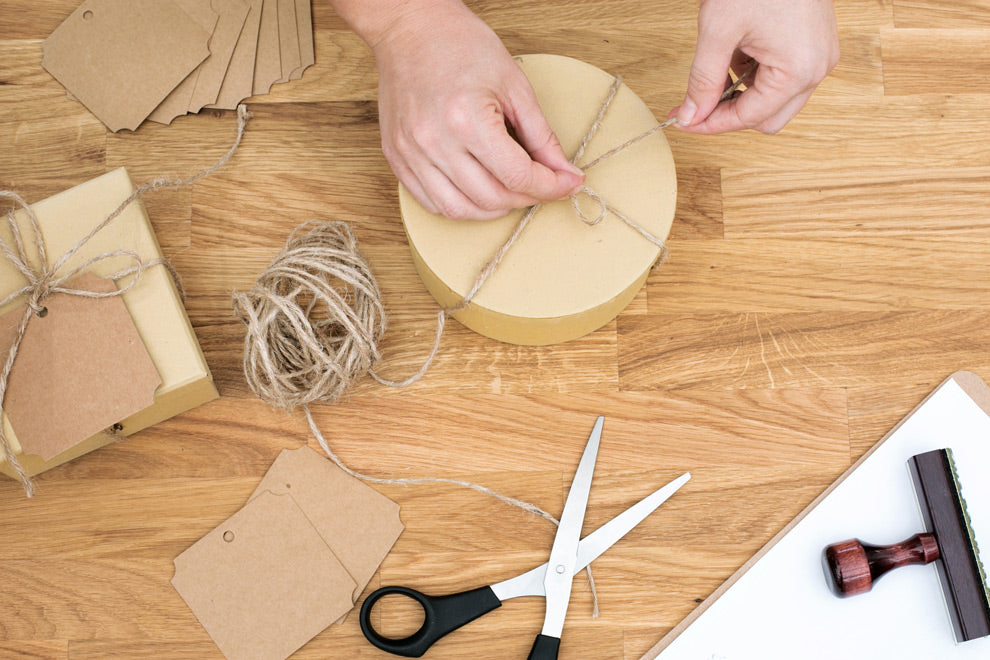 Hände binden natürliche Kordel um eine minimalistische Verpackung aus Karton auf einem hellen Holztisch neben Schere und Papiermaterialien. Die warmen Naturtöne, die strukturierte Oberfläche und die handgemachte Präsentation vermitteln Sorgfalt, hochwertige Verarbeitung und eine ruhige, nachhaltige Verpackungsatmosphäre.