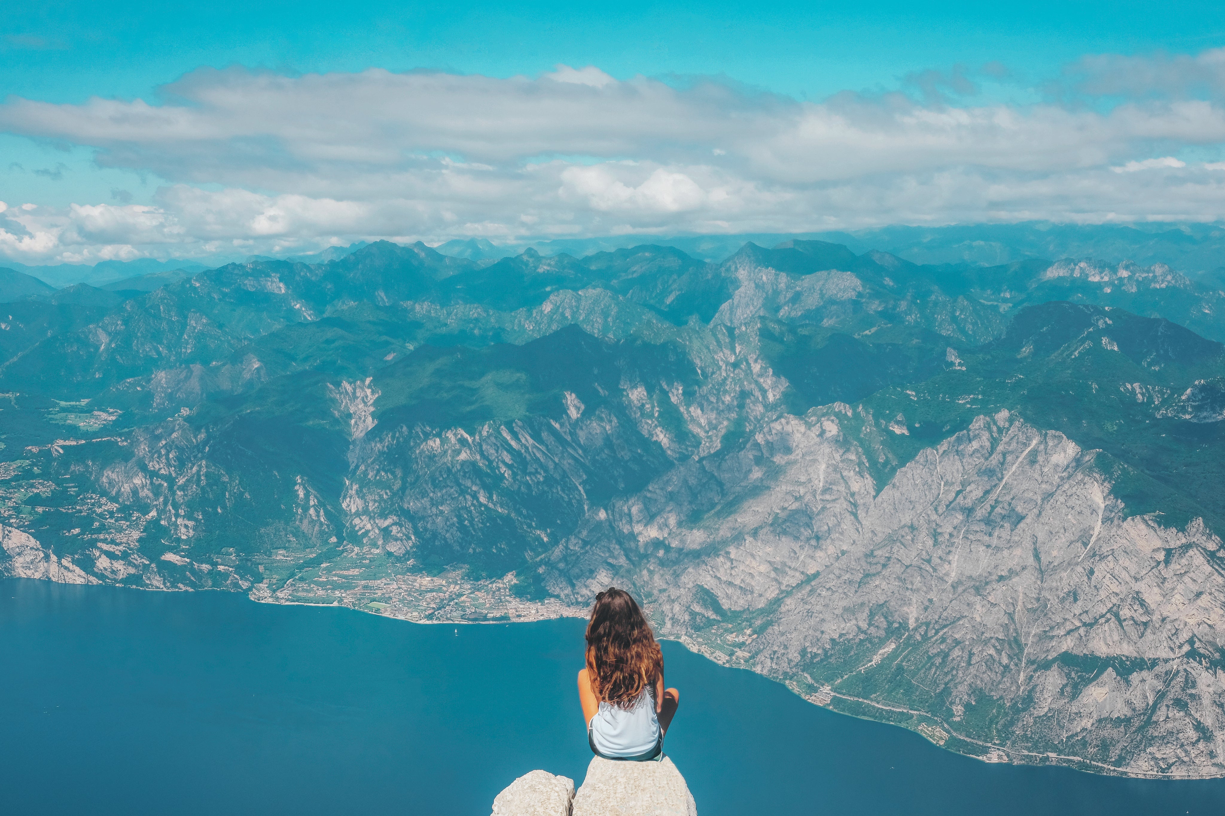 Person sitzt auf einem hellen Felsen hoch über einem tiefblauen See und blickt auf weite Berglandschaften unter einem klaren Himmel. Die Szene vermittelt frische Höhenluft, stille Ruhe und das Gefühl von Freiheit, während die kühlen Blau- und Grautöne eine entspannte, natürliche Wellness-Atmosphäre erzeugen.
