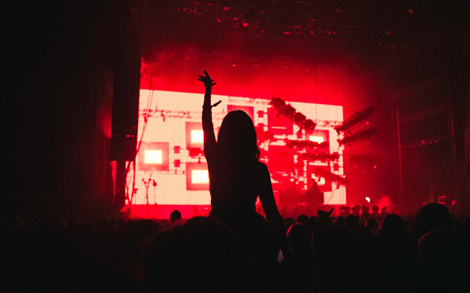 Silhouette einer Person mit erhobenem Arm vor intensiven roten Lichtflächen und unscharfen Bühnenelementen in dunkler Umgebung. Die starken Kontraste, die dynamische Lichtstimmung und die moderne Event-Atmosphäre unterstützen die visuelle Darstellung von Energie, Bewegung und urbanem Lifestyle.