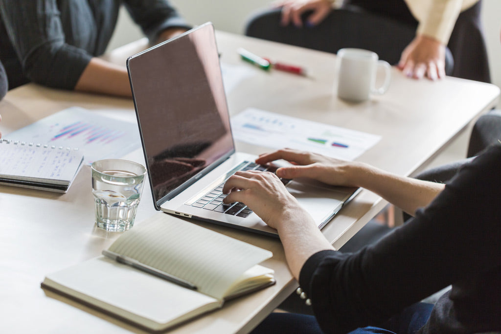 Mehrere Personen arbeiten gemeinsam an einem Tisch mit Laptop, Notizbuch, Unterlagen und Getränken in moderner Büroumgebung. Helle Farben, natürliche Lichtstimmung und die aufgeräumte Arbeitsfläche vermitteln eine strukturierte und konzentrierte Alltagssituation.